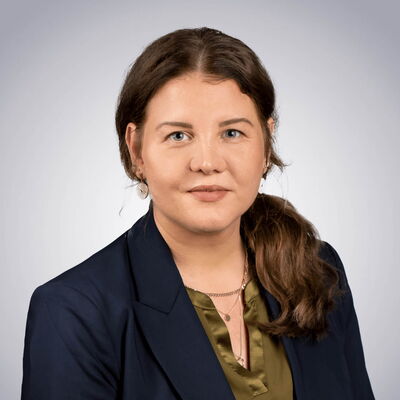 Eine Frau mit braunem Haar und blauem Blazer lächelt vor einem neutralen Hintergrund in die Kamera.