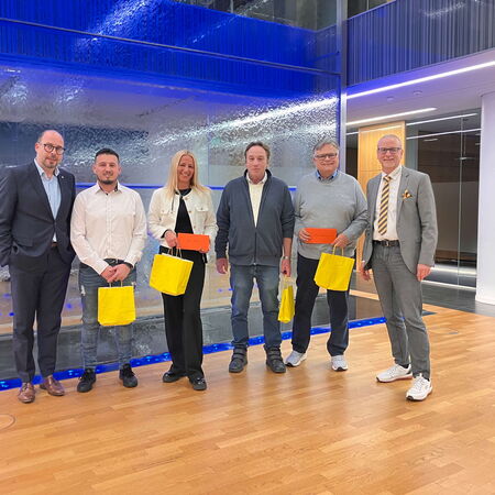 Sechs Personen stehen lächelnd in einem modernen Raum mit blauen Lichtern und tragen gelbe Geschenktüten.
