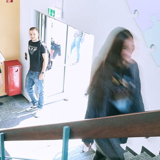 Eine Frau geht die Treppe hinauf, während im Hintergrund zwei Personen vor einem Informationsbrett im IB Reutlingen stehen.