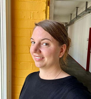 Eine lächelnde Frau steht vor einer gelben Wand in einem Flur und trägt ein schwarzes Oberteil.