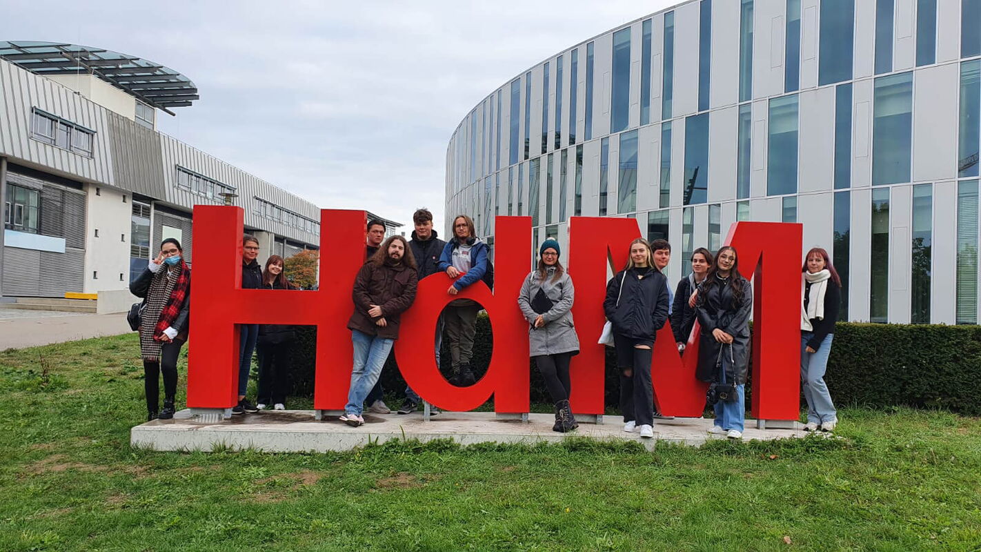 Eine Gruppe von Schülern posiert fröhlich vor einem großen roten "HdM"-Schriftzug auf einer Wiese.