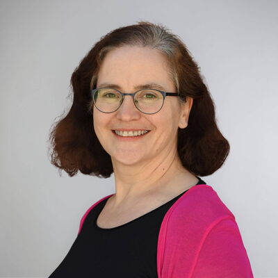 Eine lächelnde Frau mit Brille und rosa Jacke steht vor einem grauen Hintergrund.