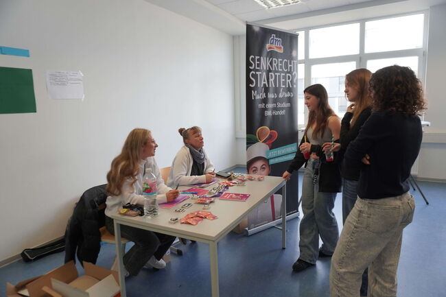Zwei Frauen informieren an einem Tisch über ein Studium im BWL-Handel, während drei Interessierte aufmerksam zuhören.