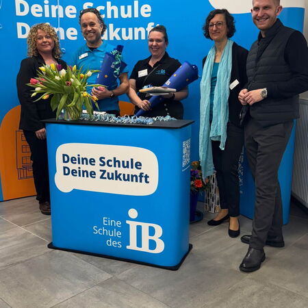 Eine Gruppe von Personen steht lächelnd vor einem blauen Banner mit dem Text "Deine Schule, Deine Zukunft" bei einer Veranstaltung.