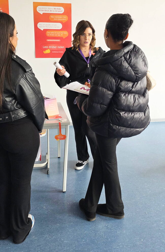 Eine Beraterin spricht mit zwei interessierten Personen über Ausbildung und Studienmöglichkeiten in einem modernen Raum.