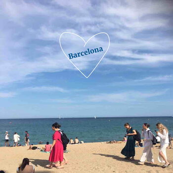 Menschen spazieren am sonnigen Strand von Barcelona unter einem blauen Himmel mit einem Herzsymbol im Hintergrund.