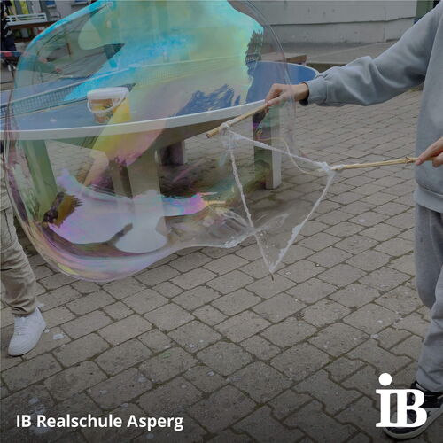 Zwei Kinder erzeugen mit Stäben und Seifenlauge eine riesige schillernde Seifenblase im Freien.