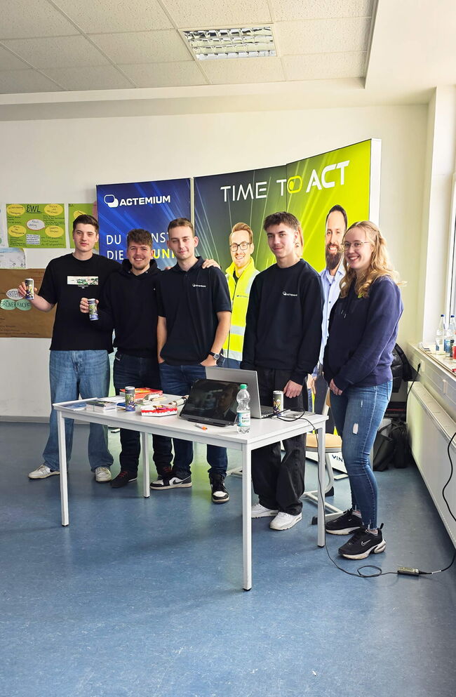 Sechs junge Menschen stehen lächelnd vor einem Tisch mit Getränken und einem Laptop, mit Actemium-Werbung im Hintergrund.