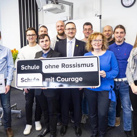 Eine Gruppe von Menschen hält ein Schild mit der Aufschrift "Schule ohne Rassismus, Schule mit Courage" in einem Raum.