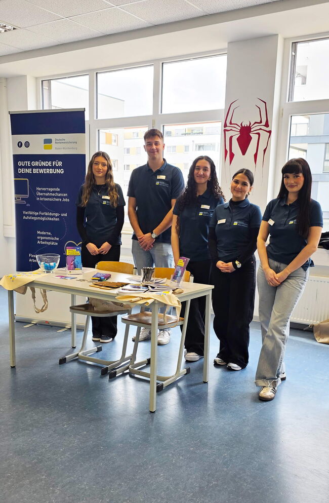 Eine Gruppe von fünf Personen steht an einem Tisch mit Informationsmaterialien für Bewerbungen in der Berufswelt.