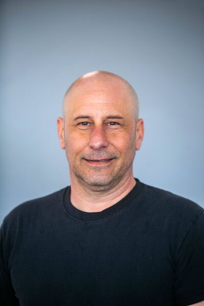 Ein Mann in einem schwarzen T-Shirt lächelt vor einem neutralen, grauen Hintergrund.