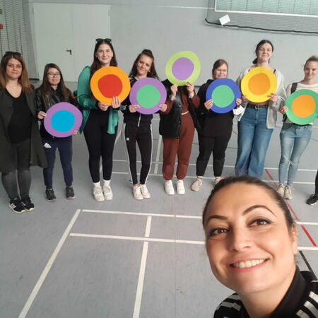 Eine Gruppe von Menschen hält bunte Kreise in einer Sporthalle, während eine Frau vorne ein Selfie macht.