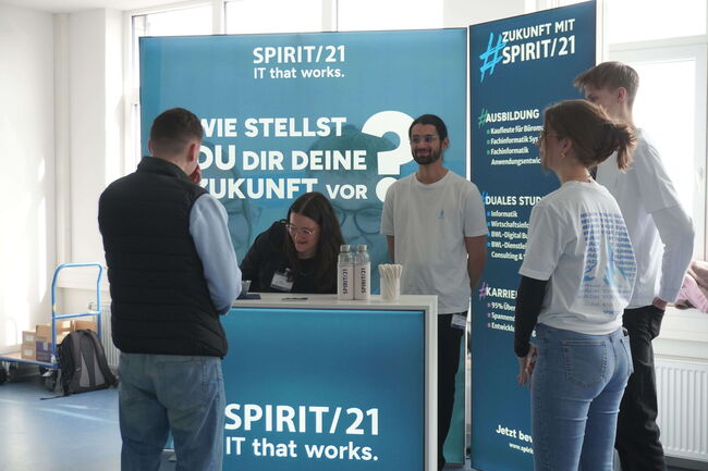 Mehrere Menschen stehen an einem Messestand von SPIRIT/21 und diskutieren über IT-Karrieremöglichkeiten.