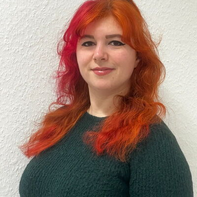 Eine Frau mit leuchtend orange-rotem Haar lächelt in einem dunkelgrünen Pullover vor einer weißen Wand.