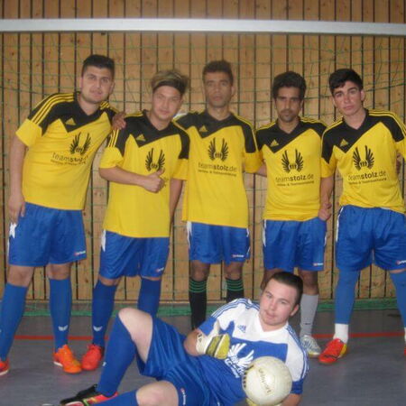 Eine Gruppe junger Männer in gelben Trikots posiert in einer Sporthalle mit einem liegenden Torwart und einem Fußball.