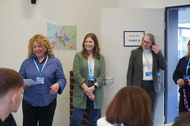 Drei Frauen stehen lächelnd in einem Klassenzimmer mit einer Europakarte an der Wand und führen eine Präsentation durch.