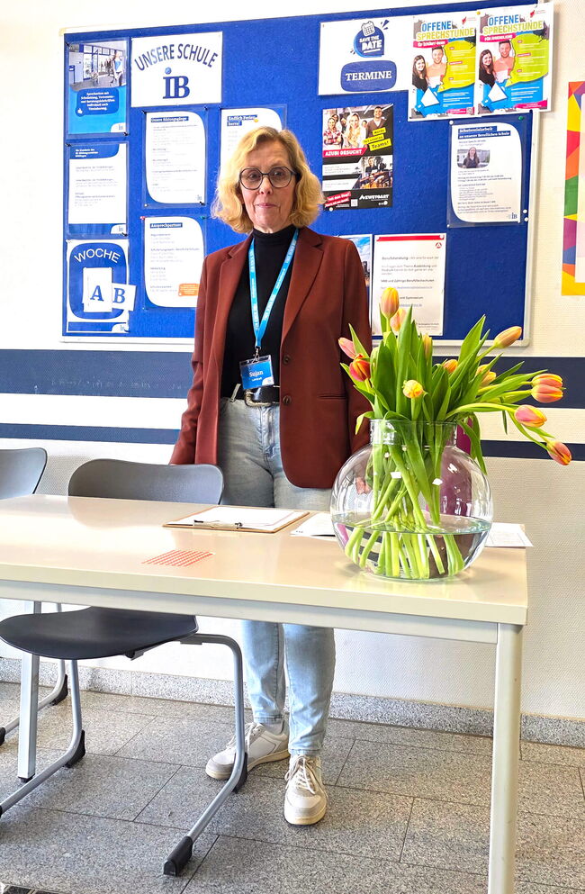 Eine Lehrkraft steht an einem Tisch mit einer Vase voll Tulpen, während im Hintergrund Informationen zur Schule präsentiert werden.