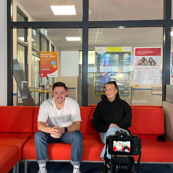 Zwei Personen sitzen auf einem roten Sofa und werden für ein Interview von einer Kamera aufgenommen.