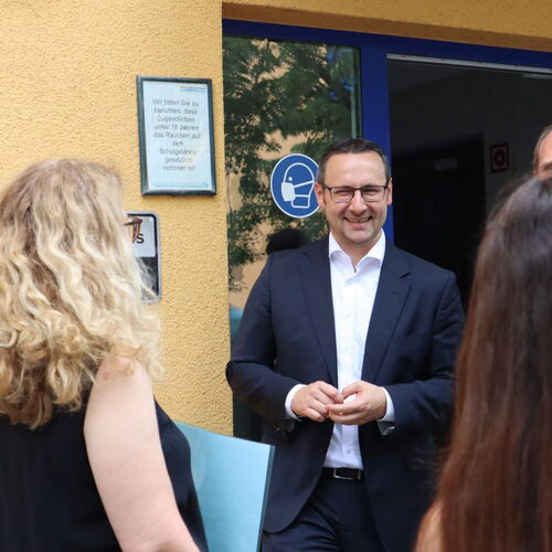 Eine Gruppe von Menschen unterhält sich vor einem Schulgebäude, während ein Mann im Anzug lächelnd zuhört.