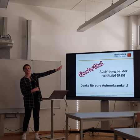 Eine Person präsentiert in einem Raum vor einem Bildschirm, der Informationen zur Ausbildung bei HERRLINGER KG zeigt.