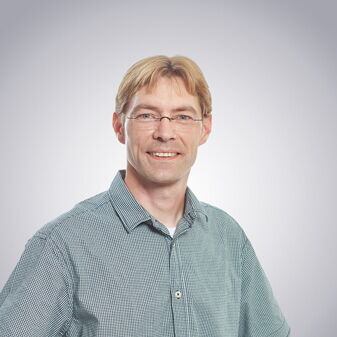 Ein lächelnder Mann mit Brille und kariertem Hemd steht vor einem neutralen Hintergrund in Stuttgart-Mitte.