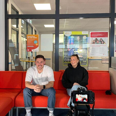 Zwei Personen sitzen auf einem roten Sofa und werden für ein Interview von einer Kamera aufgenommen.