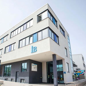 Moderne Außenansicht eines weißen Gebäudes mit großen Fenstern und einem "IB"-Logo an der Fassade.