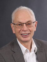 Ein lächelnder älterer Mann mit Brille trägt einen grauen Anzug und ein weißes Hemd vor einem neutralen Hintergrund.
