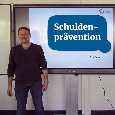 Ein Mann steht lächelnd neben einem Bildschirm, der das Thema "Schuldenprävention" präsentiert.
