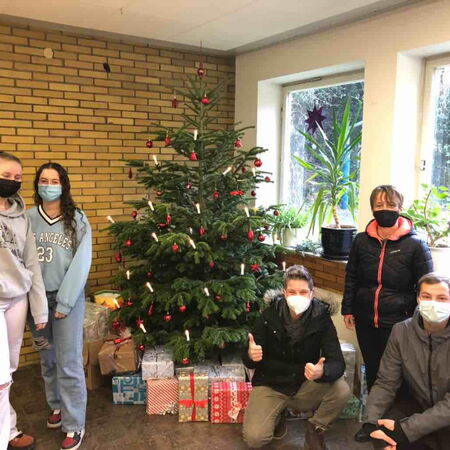 Fünf Personen mit Masken posieren lächelnd neben einem festlich geschmückten Weihnachtsbaum und zahlreichen Geschenken.