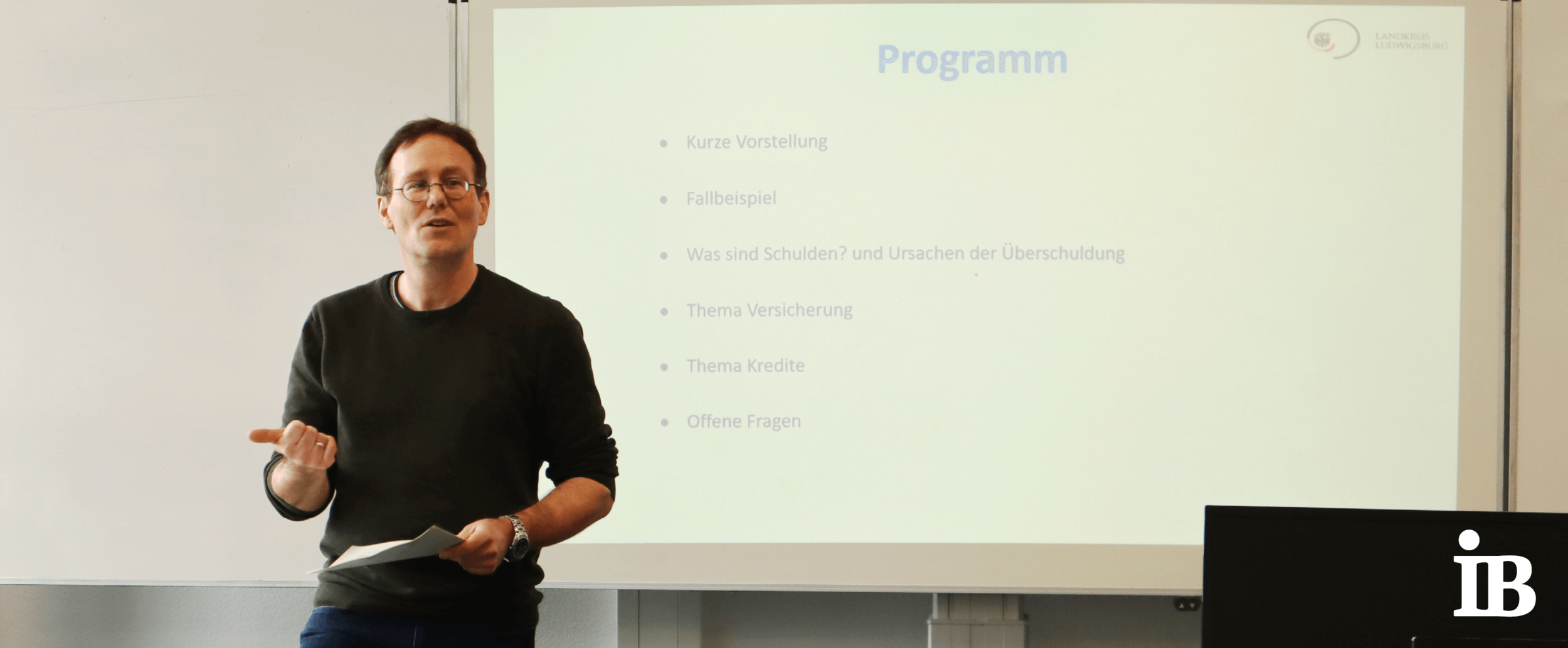 Ein Mann hält eine Präsentation über Schuldenprävention vor einem Programmplan auf einer Leinwand.