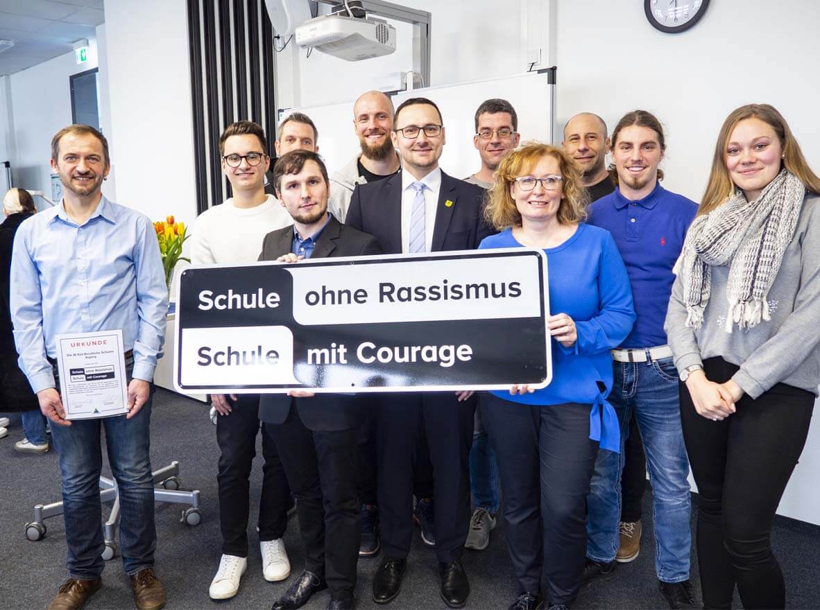 Eine Gruppe von Menschen hält ein Schild mit der Aufschrift "Schule ohne Rassismus, Schule mit Courage" in einem Raum.