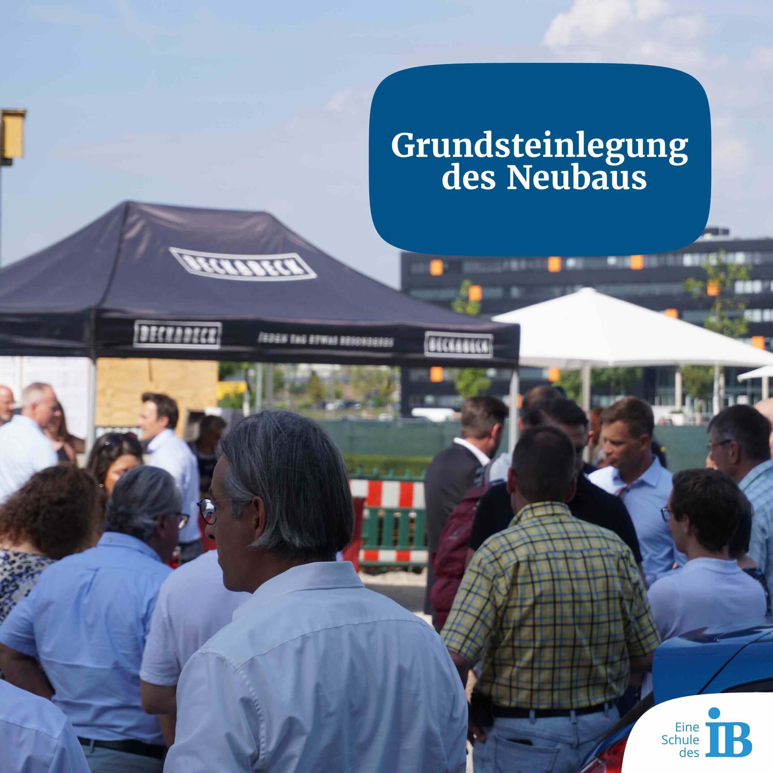 Menschen versammeln sich bei sonnigem Wetter zur Grundsteinlegung eines Neubaus unter einem Zelt.