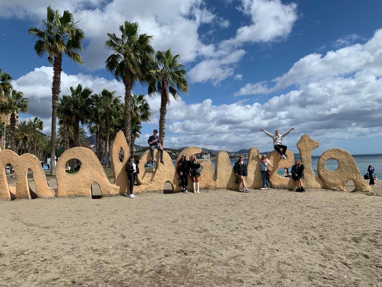 Menschen posieren fröhlich auf dem großen "Malagueta"-Schriftzug am Strand von Málaga unter Palmen und blauem Himmel.