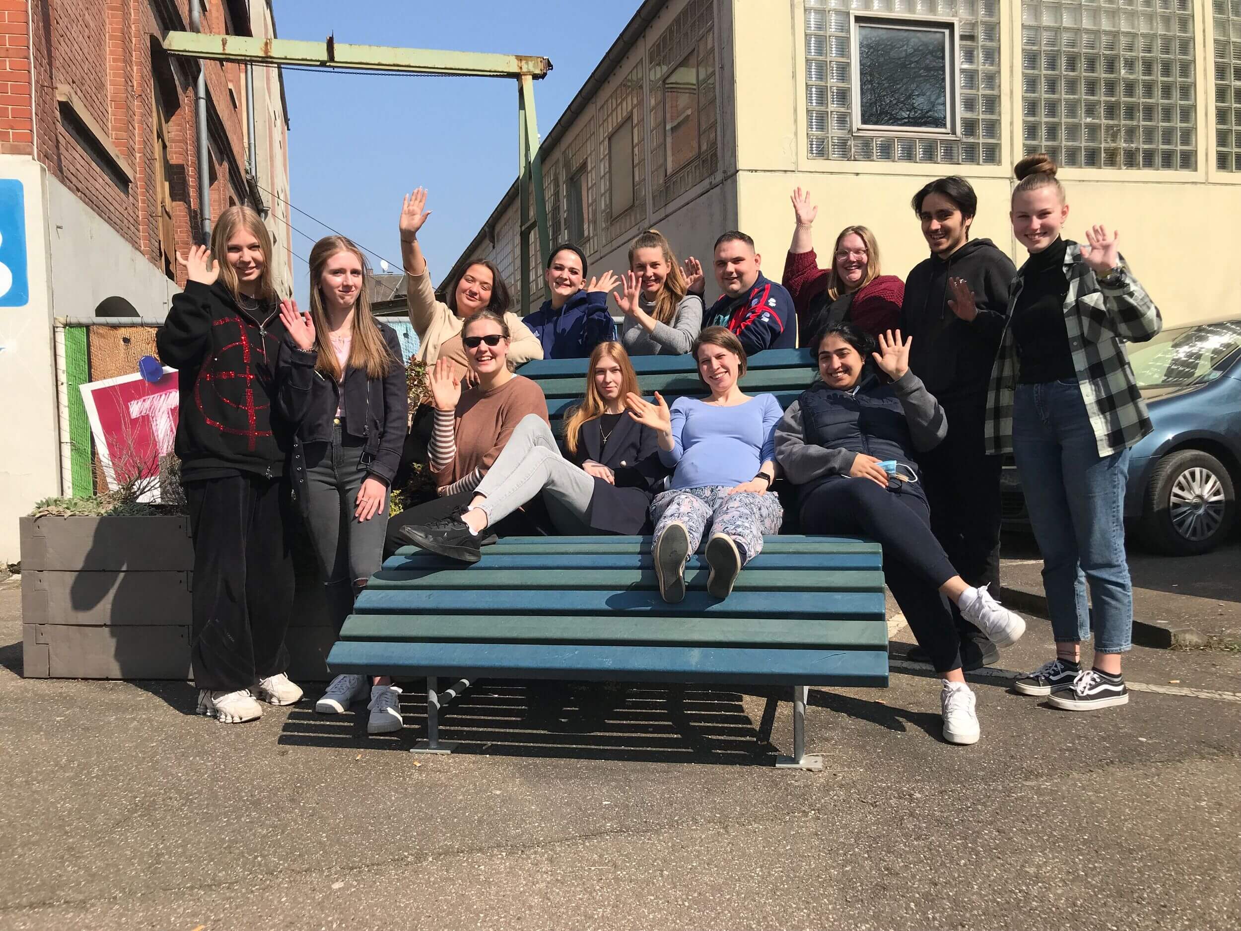 Eine Gruppe von Menschen sitzt auf einer großen Bank im Freien und winkt fröhlich in die Kamera.
