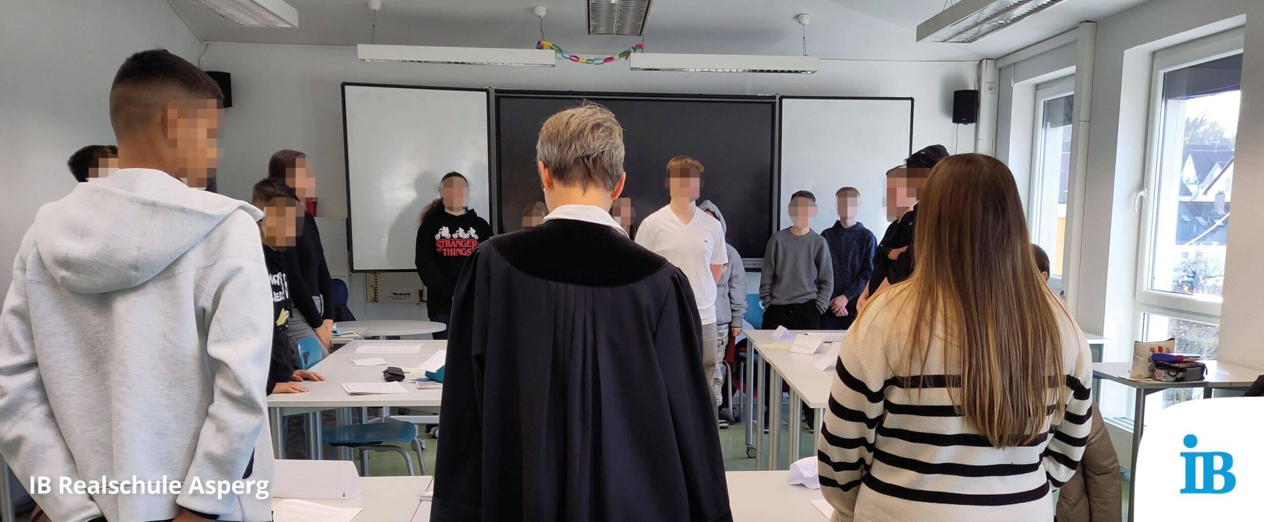 Eine Gruppe von Schülern steht in einem Klassenzimmer um einen Lehrer im Talar, der vor einem Whiteboard spricht.