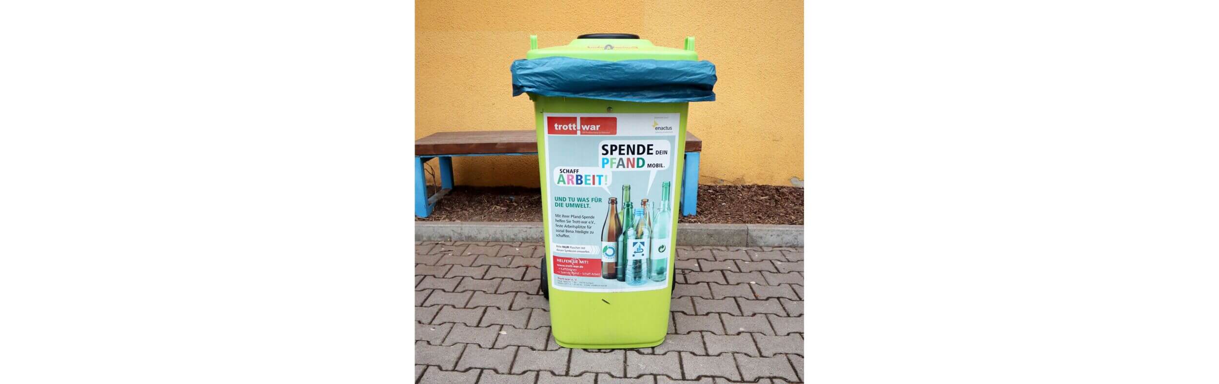 Eine gelbe Pfandtonne steht vor einer Schule mit einem Plakat, das zur Spende von Pfandflaschen aufruft.