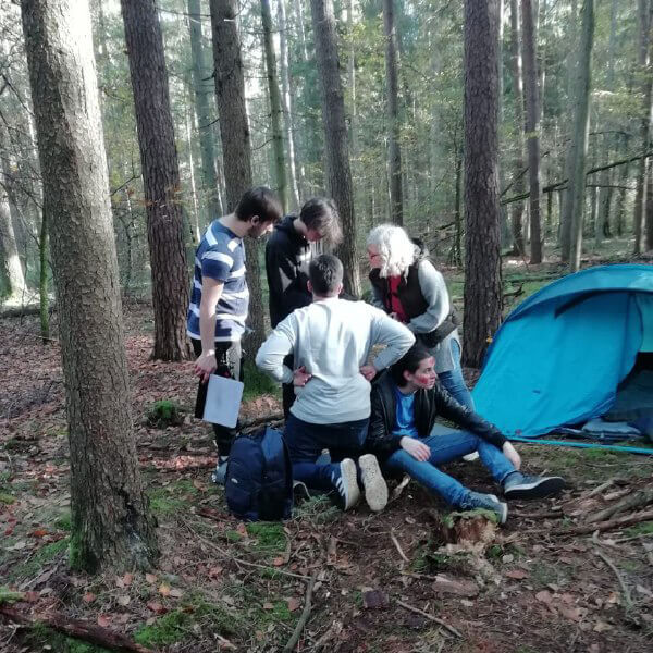 Eine Gruppe von Menschen steht und kniet um ein Lagerfeuer im Wald neben einem blauen Zelt bei Ludwigswinkel.