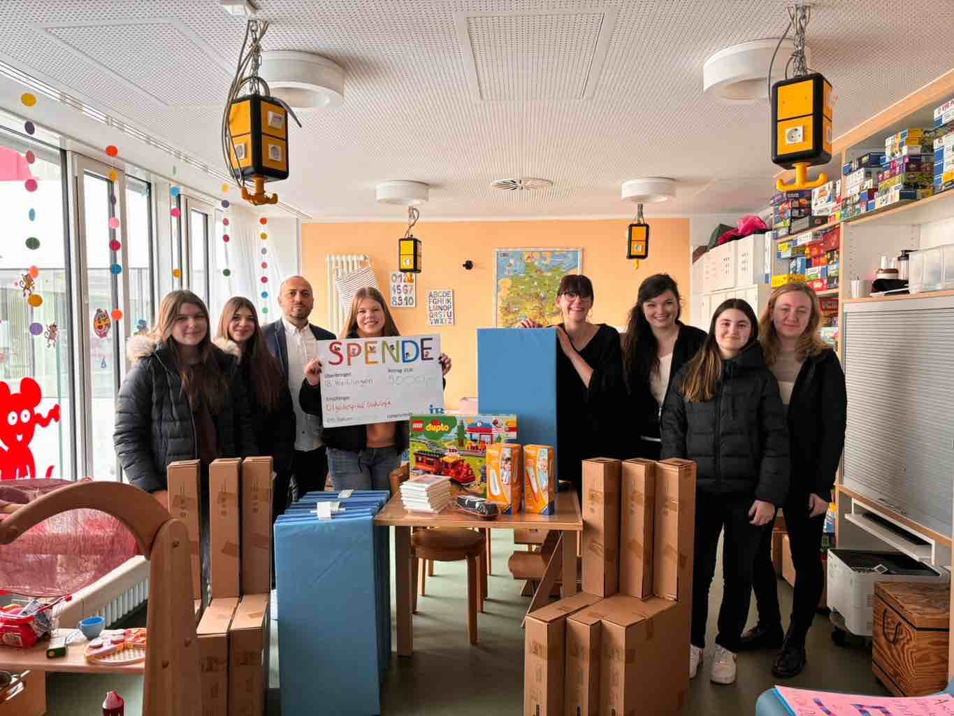 Eine Gruppe von Menschen steht in einem Raum mit Spielsachen und hält ein großes Spendenplakat für wohltätige Zwecke.