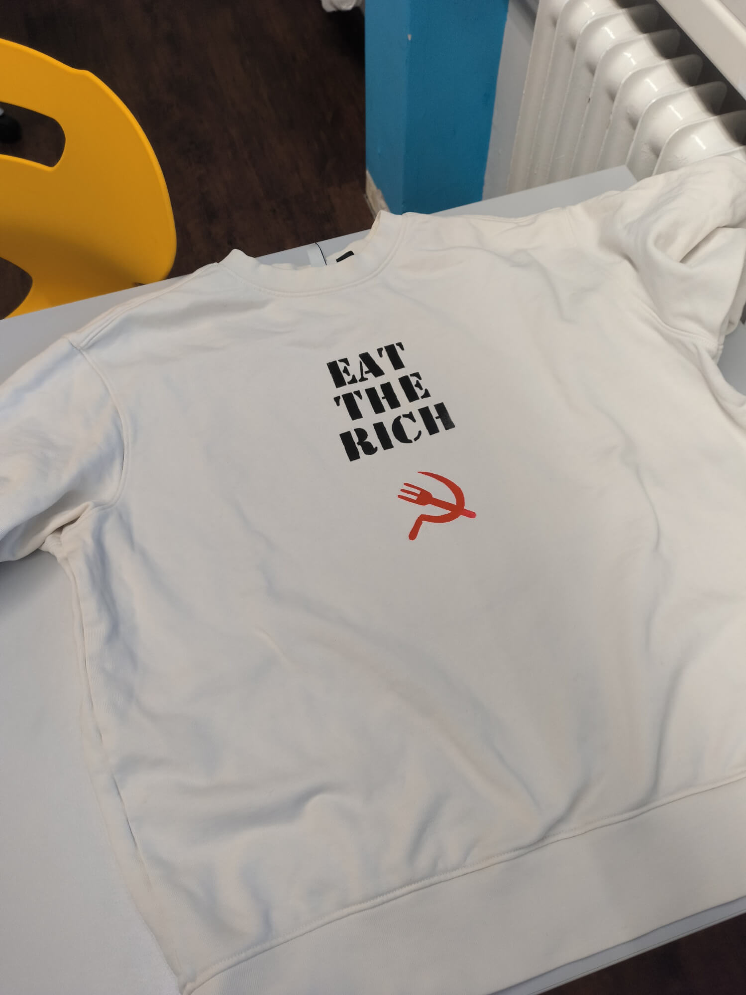 Ein weißer Pullover liegt auf einem Tisch und zeigt den Aufdruck "EAT THE RICH" mit einer roten Gabel.
