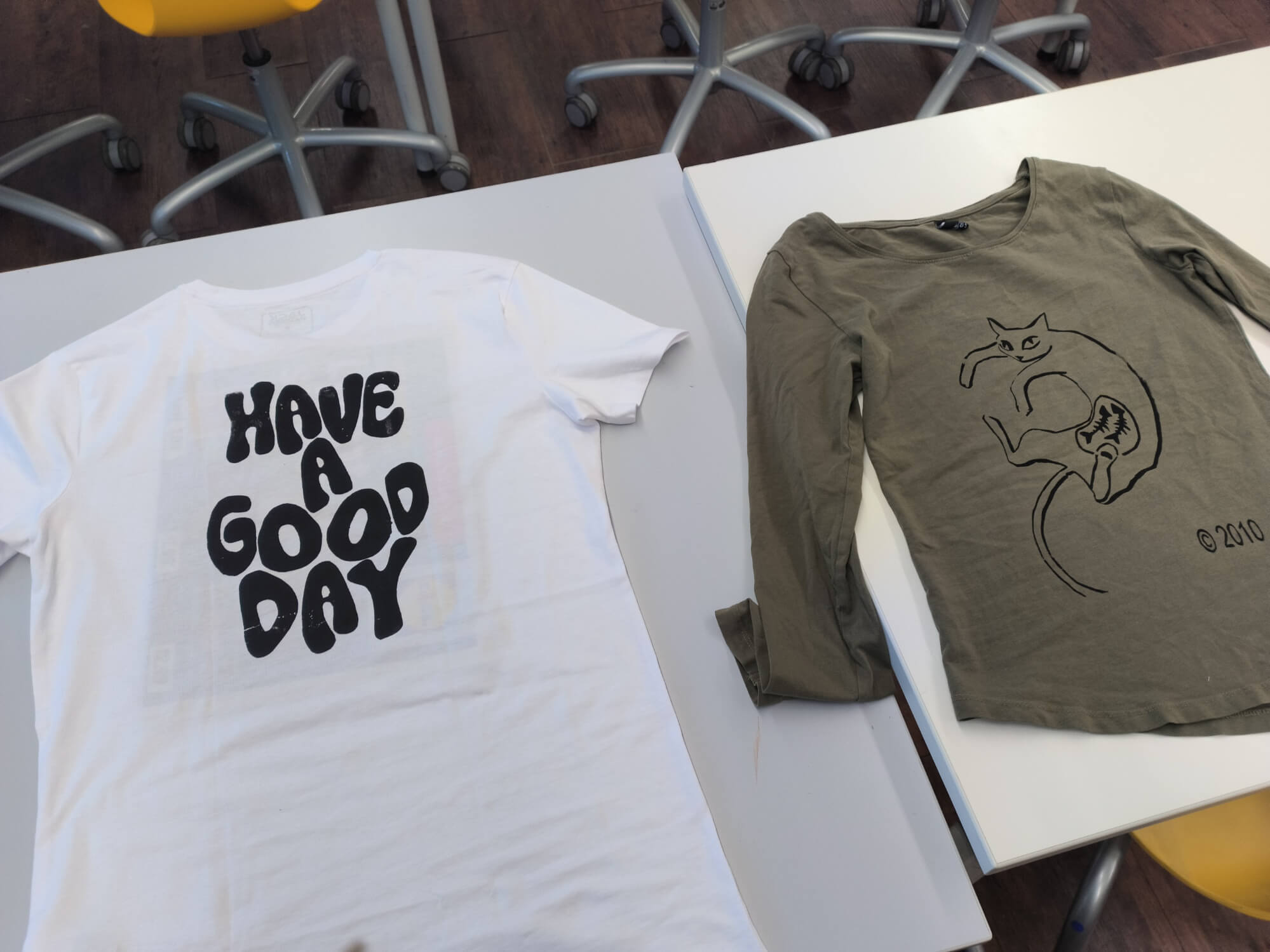 Zwei T-Shirts liegen auf einem Tisch, eines mit dem Aufdruck "Have a Good Day" und das andere mit einer Katze.