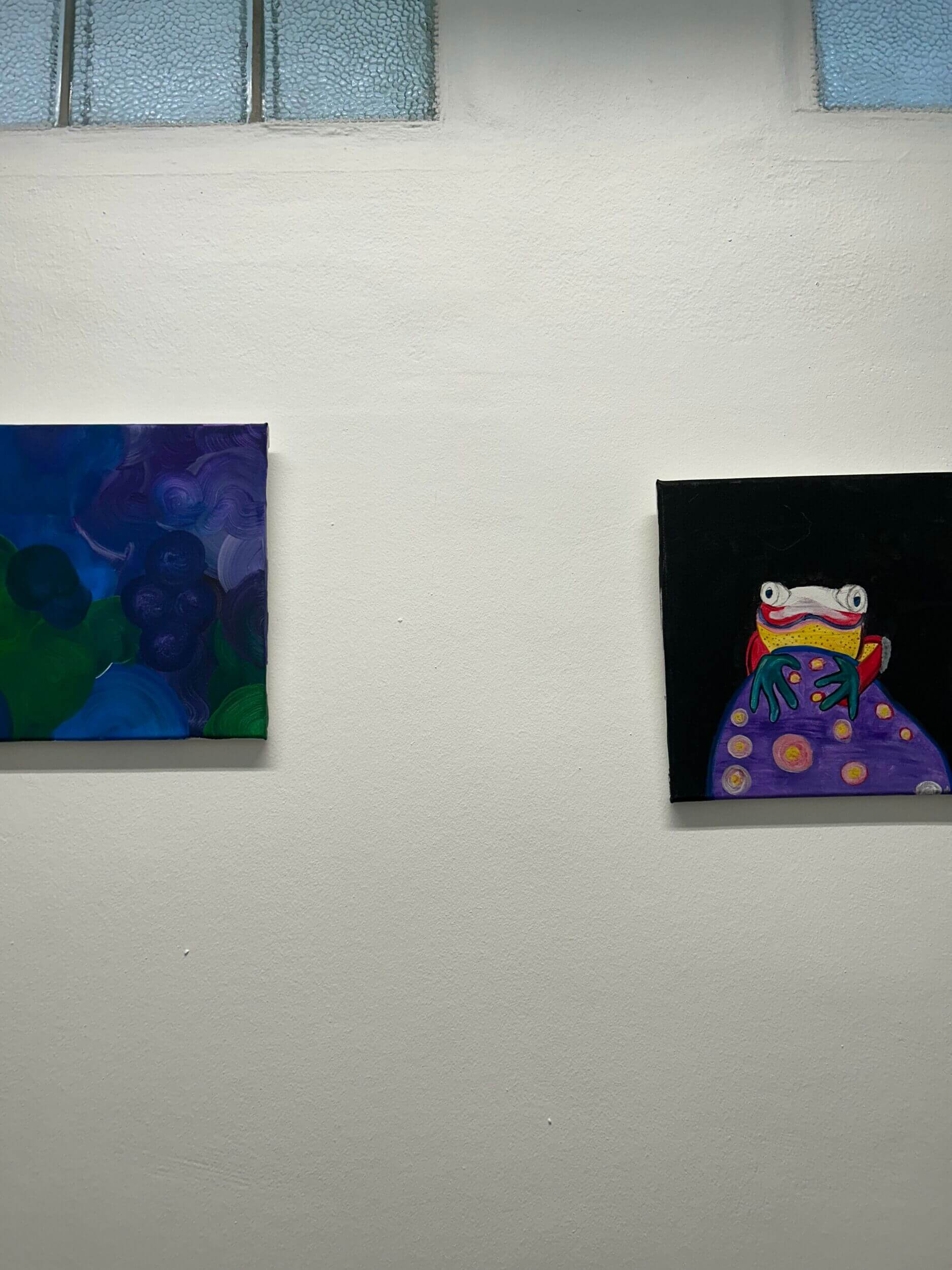 Zwei bunte Gemälde hängen an einer weißen Wand, eines zeigt einen farbenfrohen Frosch.