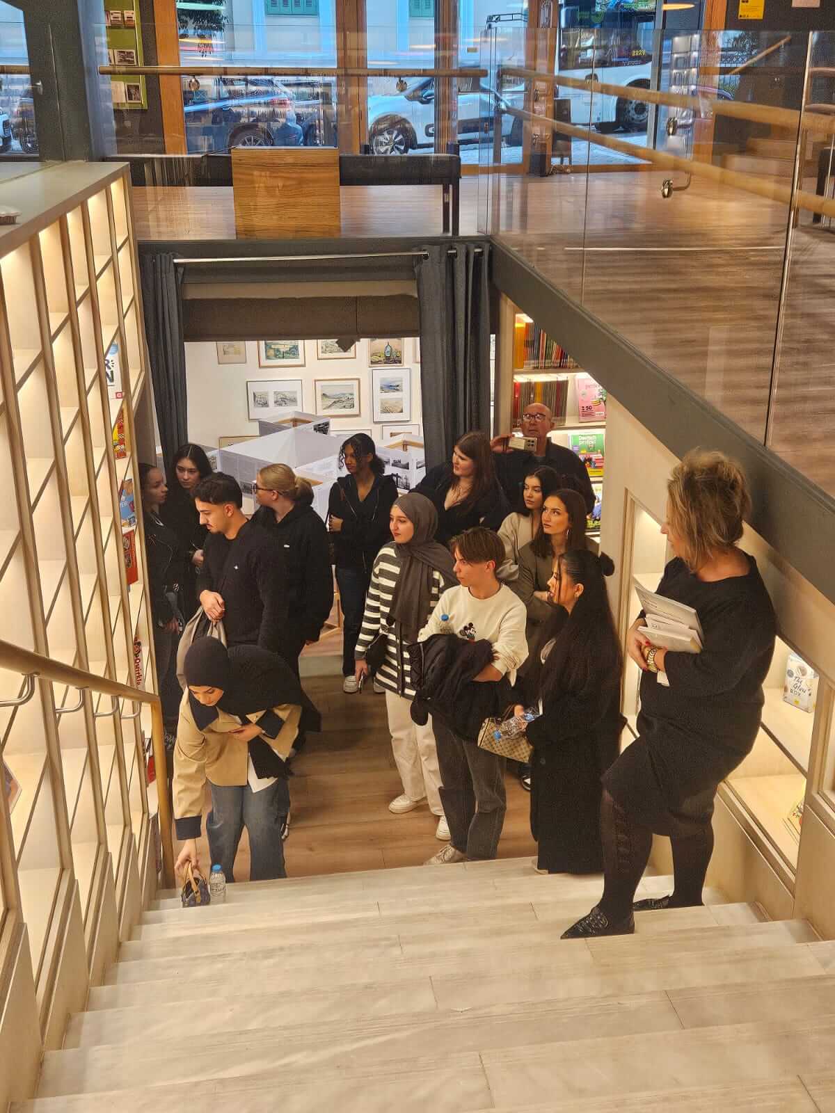 Eine Gruppe von Menschen versammelt sich auf einer Treppe in einem modernen Gebäude, während eine Frau sich nach einem Gegenstand bückt.