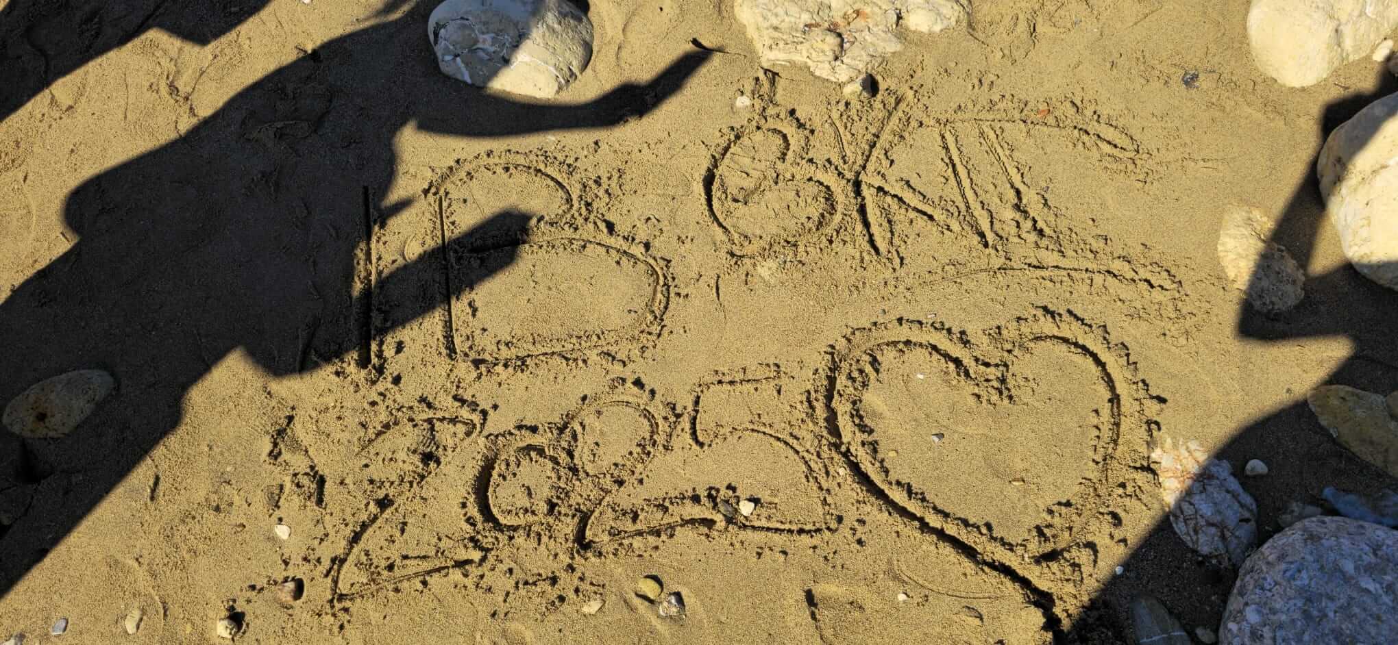 Auf dem Sand steht "B 2025" und ein Herz, umgeben von Schatten und Steinen.