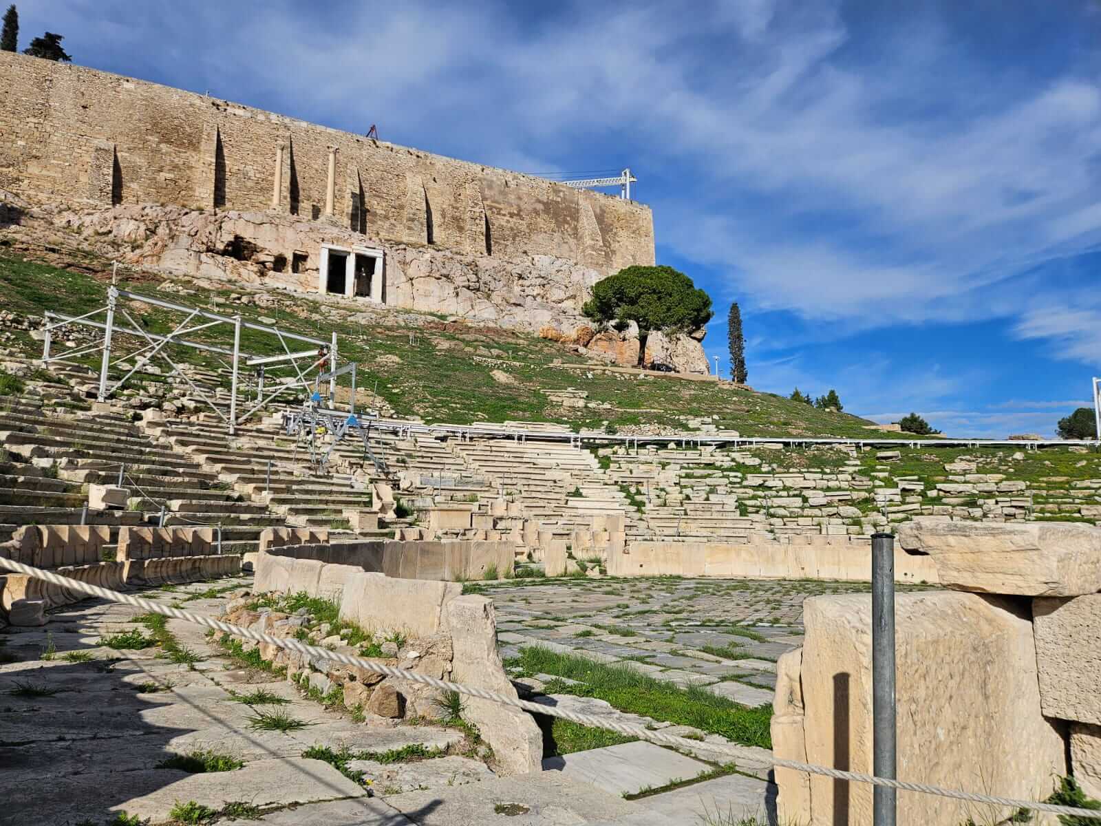 Das antike Amphitheater mit steinernen Sitzreihen steht unter einem klaren blauen Himmel, während Baugerüste sichtbar sind.