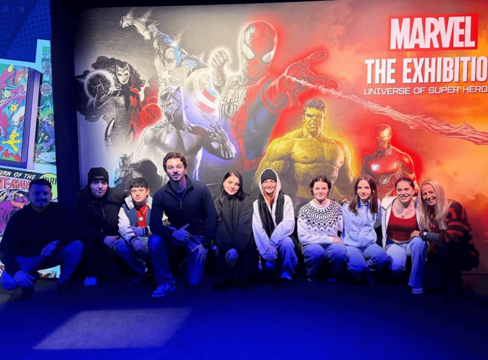 Eine Gruppe von Menschen posiert vor einem Marvel-Poster mit Superhelden bei einer Ausstellung.