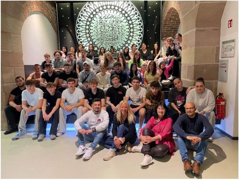 Eine große Gruppe von Menschen posiert vor einem kunstvollen QR-Code-Muster in einem Gebäude in Nürnberg.
