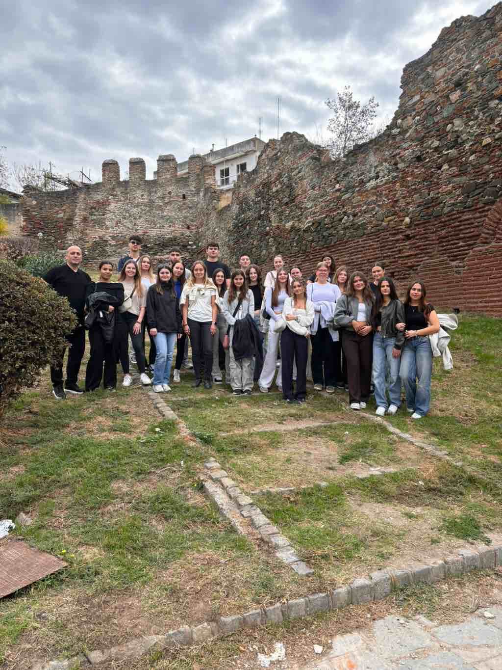 Eine Gruppe von Menschen posiert vor einer antiken Mauer in Thessaloniki unter bewölktem Himmel.