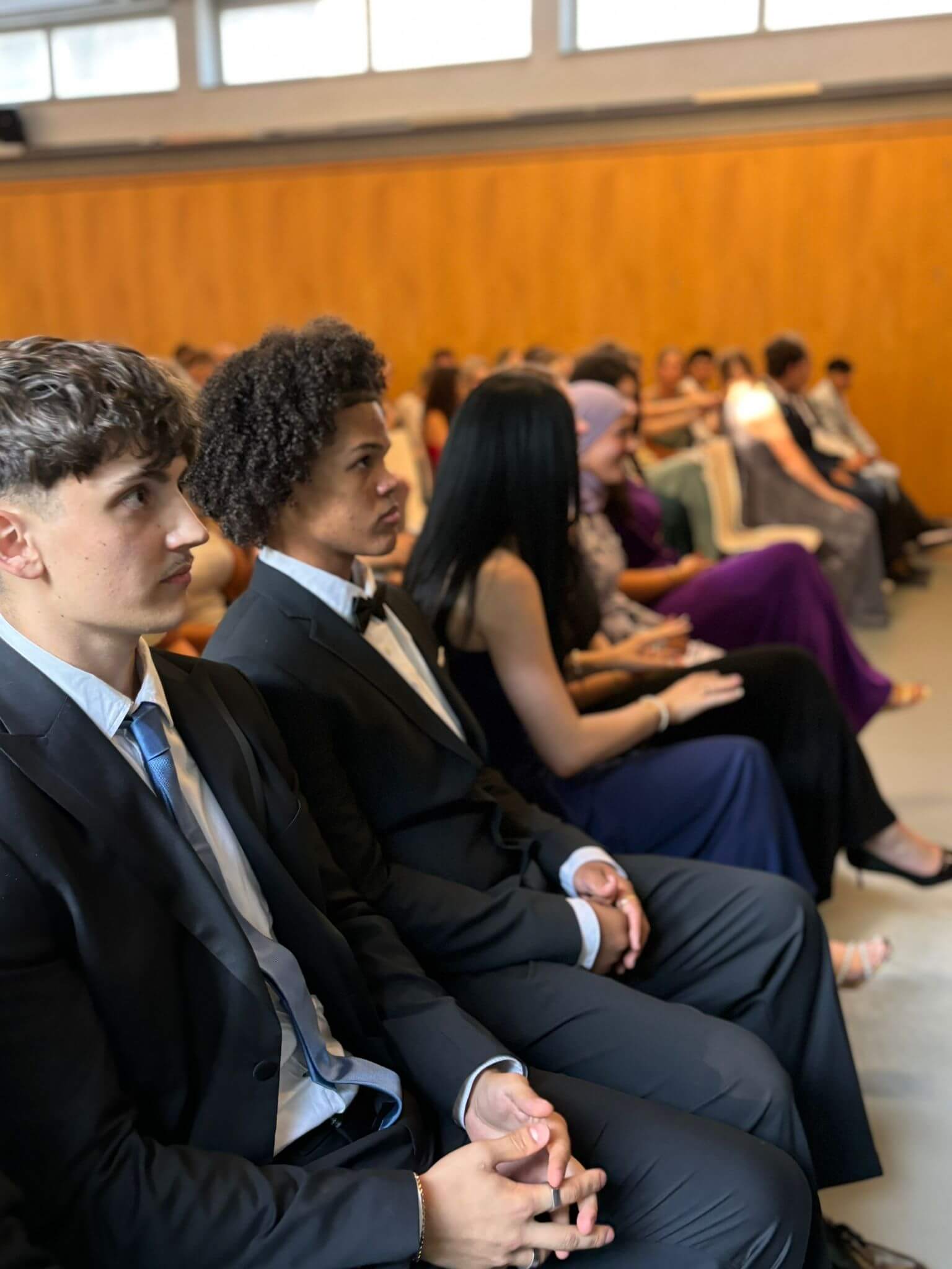 Zwei junge Männer in Anzügen sitzen aufmerksam in einer Reihe bei einer formellen Veranstaltung im Juli 2025.
