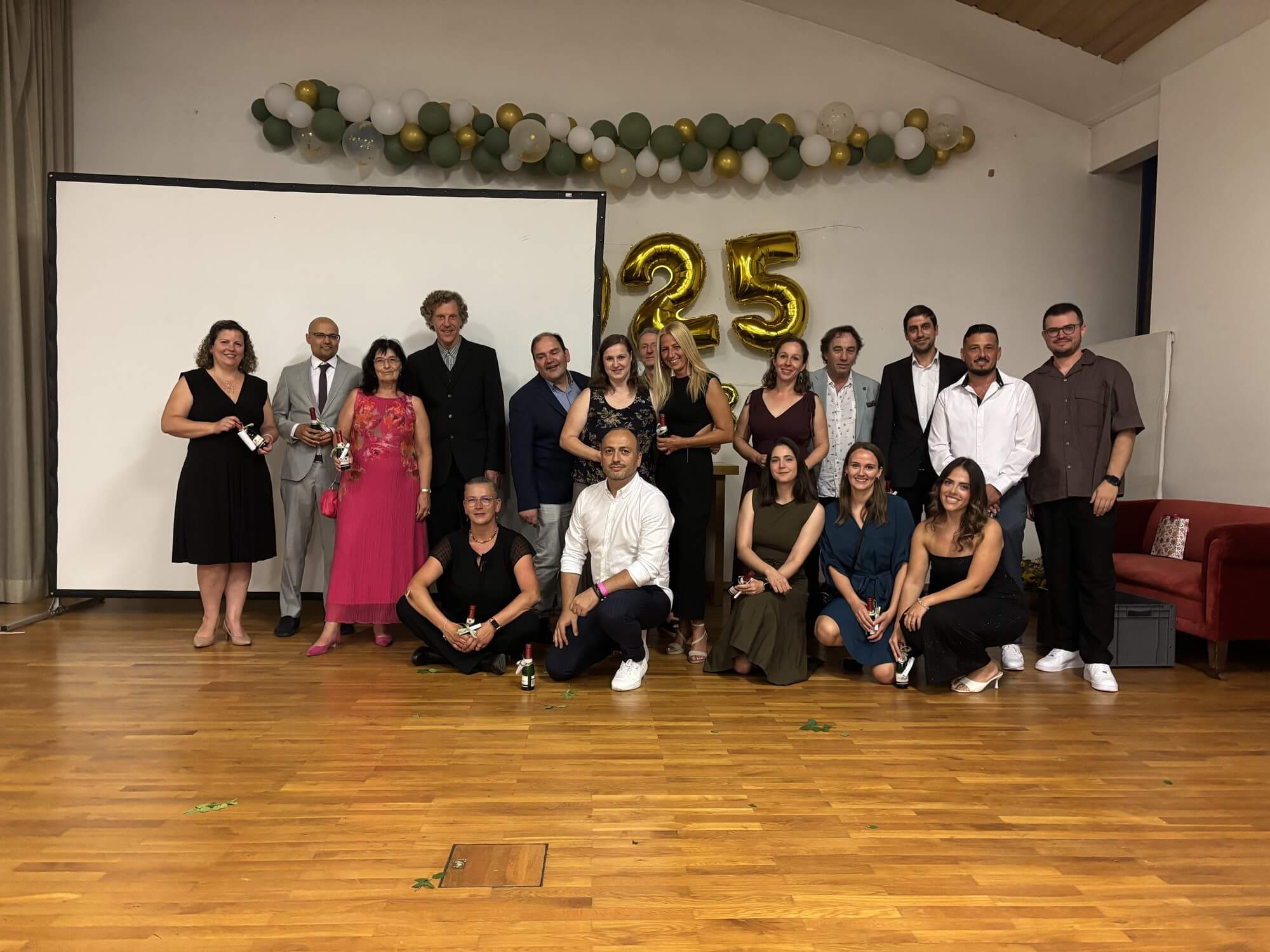 Eine Gruppe von Menschen feiert ein 25-jähriges Jubiläum vor goldenen Ballons in einem festlich geschmückten Raum.